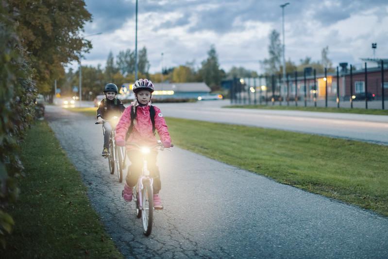 Kaksi kypäräpäistä lasta pyöräilee hämärällä valot loistaen.