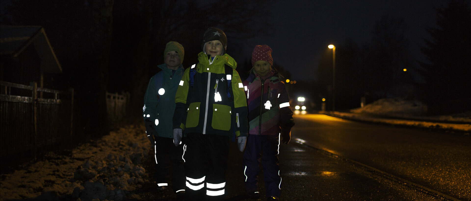 Kolme lasta kävelee pimeässä heijastimet loistaen.
